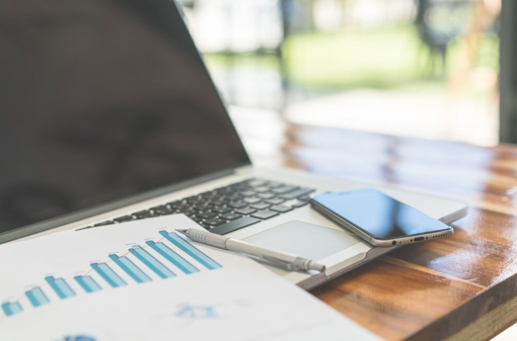 A blurred laptop, phone, and printed graphs on a wooden table suggesting remote work