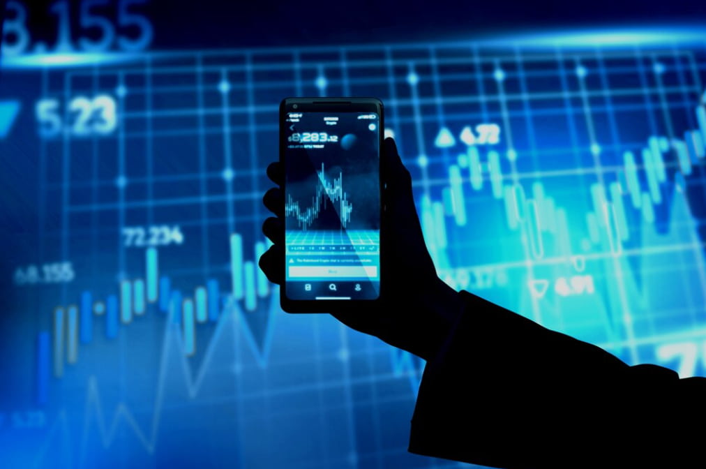 A hand holding a smartphone with stock market data on screen against a digital graph background