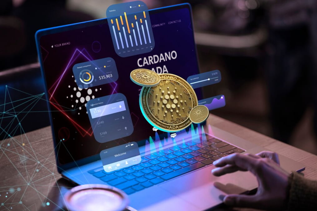 an opened laptop and hand typing on the touch bar, Cardano coins, and trading icons near the screen