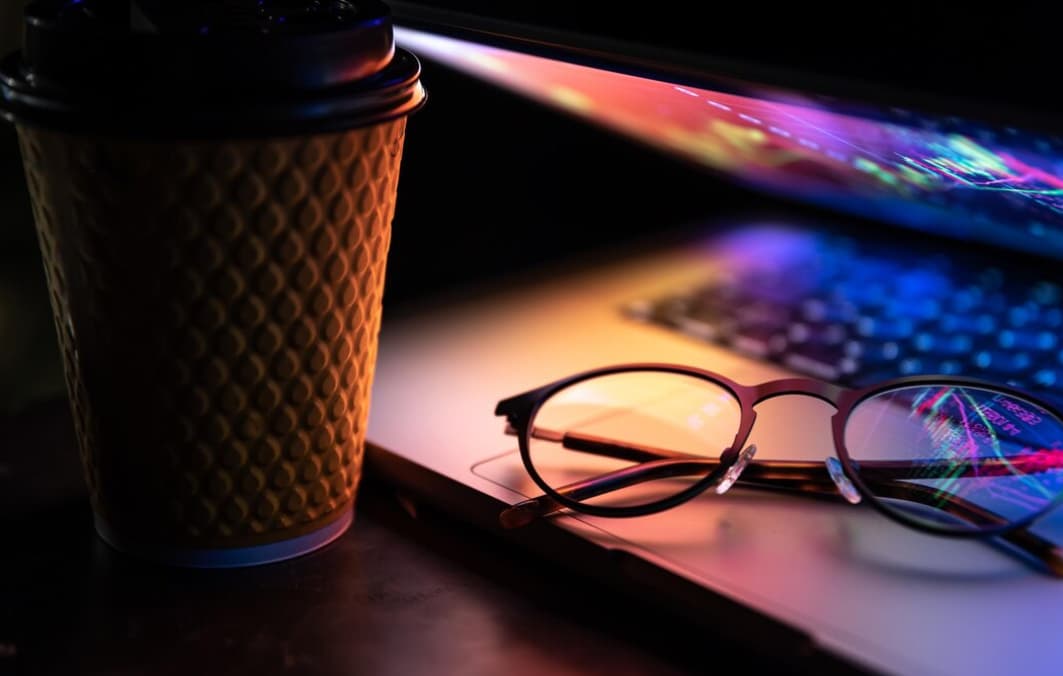 A coffee cup and glasses on a laptop keyboard in low light