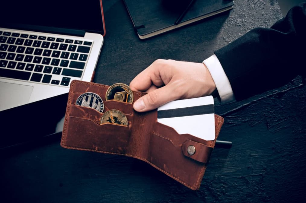 hand holds brown leather wallet with card and three coins inside, a laptop and notebook on the table